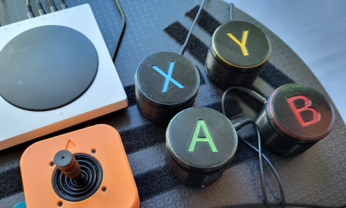 A photo of 4 assistive switches, a joystick, and an Xbox Adapitve Controller on a laptop tray with velcro.