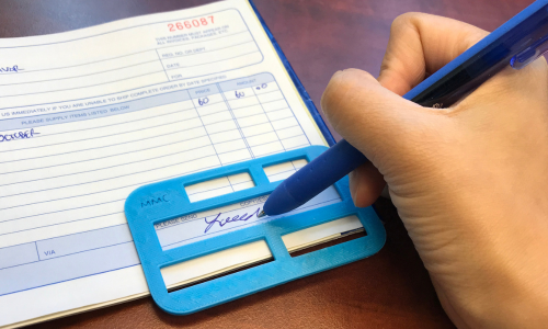 A close up shot of someones hand using a pen and MMC Signature Guide device to sign up on a page.