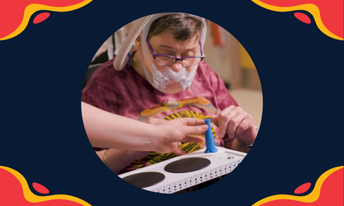 A graphic with red and yellow blobs in all 4 corners of the rectangle with a circular photo in the center showing a person with a disability looking at a 3D printed joystick on an Xbox Adaptive Controller. A hand of another person coming from the side of the picture is pointing to the joystick.