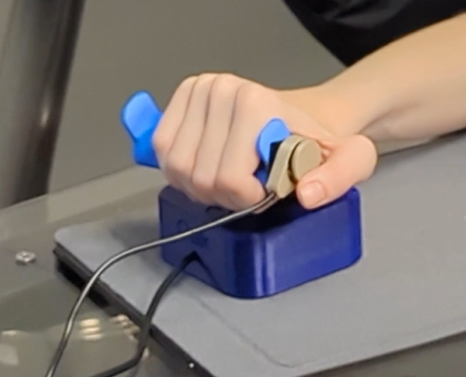 a hand on an oak joystick with a goalpost topper and a blue raindrop assistive switch mounted on the side arm of the goalpost topper and the users thumb hovering over it to press it